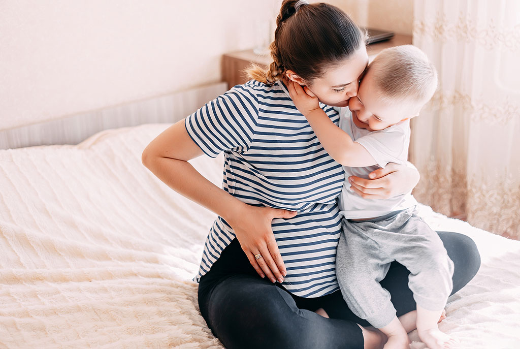 A Pregnant Mother Sitting on a Bed Hugging Her Other Child What Is Secondary Infertility